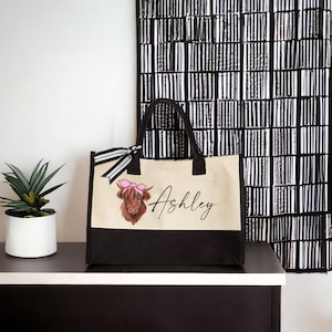 K&ouml;nnte beinhalten: Eine beige und schwarze Canvas-Tragetasche mit einer braunen Highland-Kuh mit einer rosa Schleife auf dem Kopf. Die Tasche hat einen schwarzen Griff und der Name "Ashley" ist auf der Vorderseite aufgedruckt.