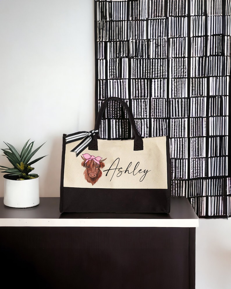 K&ouml;nnte beinhalten: Eine beige und schwarze Canvas-Tragetasche mit einer braunen Highland-Kuh mit einer rosa Schleife auf dem Kopf. Die Tasche hat einen schwarzen Griff und der Name "Ashley" ist auf der Vorderseite aufgedruckt.