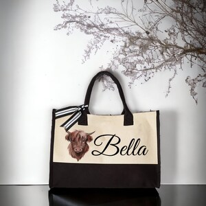 K&ouml;nnte beinhalten: Eine beige und braune Canvas-Tragetasche mit einem schwarz-wei&szlig; gestreiften Band. Die Tasche zeigt eine braune und wei&szlig;e Highland-Kuh-Illustration und den Namen "Bella" in schwarzer Kursivschrift.