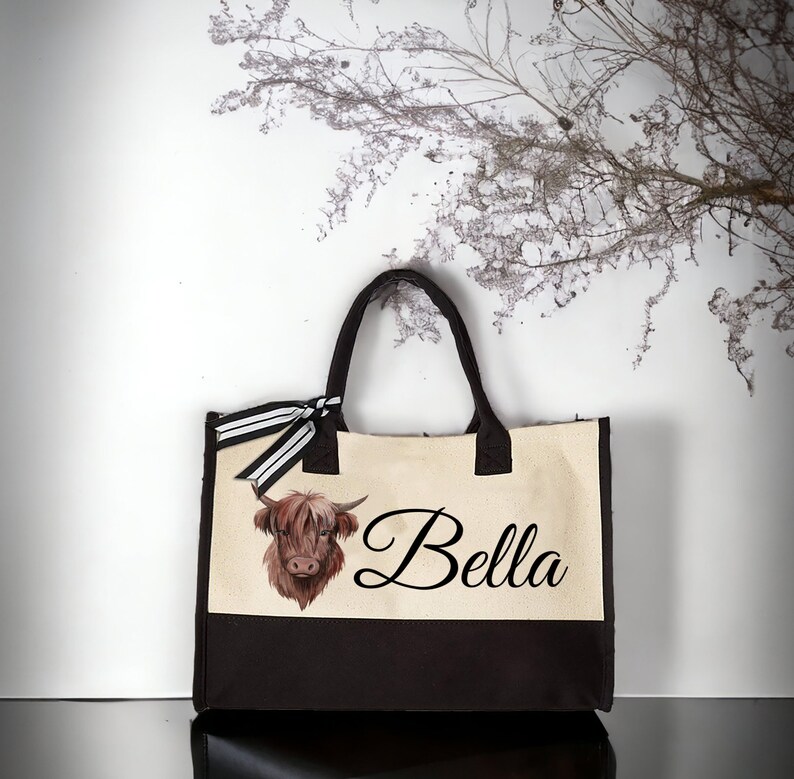 K&ouml;nnte beinhalten: Eine beige und braune Canvas-Tragetasche mit einem schwarz-wei&szlig; gestreiften Band. Die Tasche zeigt eine braune und wei&szlig;e Highland-Kuh-Illustration und den Namen "Bella" in schwarzer Kursivschrift.