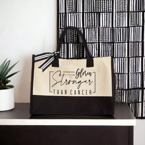 May include: A black and beige tote bag with a black ribbon and the text "Gloria Stronger Than Cancer" printed on the front.