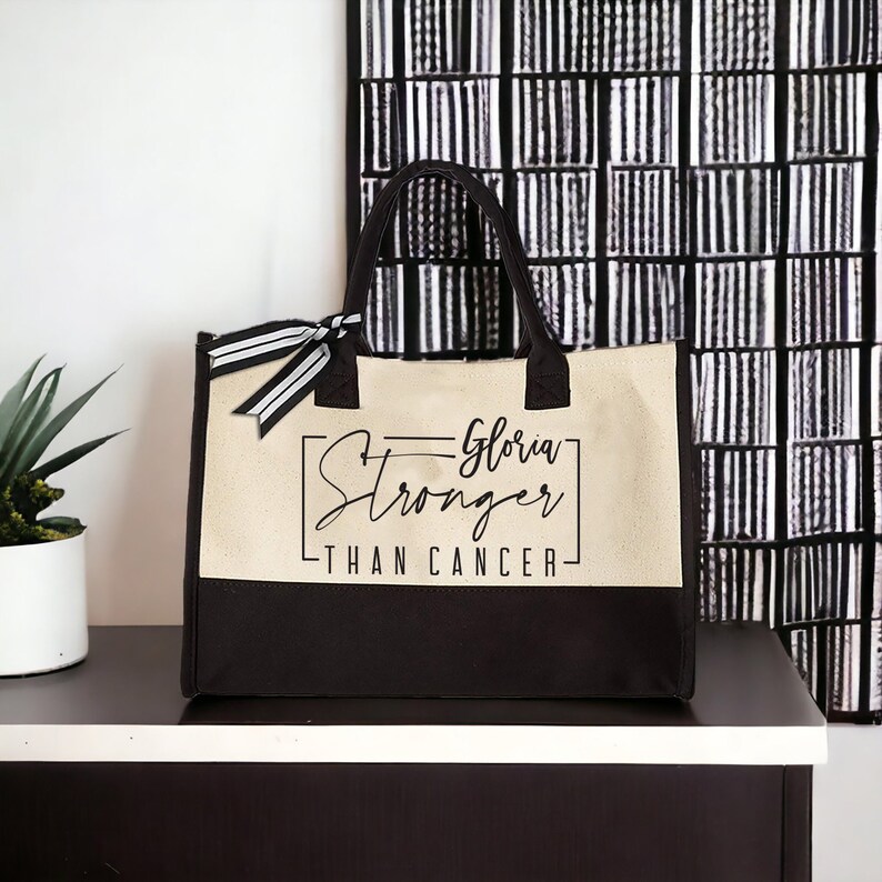 May include: A black and beige tote bag with a black ribbon and the text "Gloria Stronger Than Cancer" printed on the front.