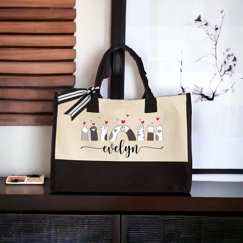 May include: A black and beige tote bag with a design of cats holding paws and hearts. The bag has a black and white striped ribbon and the name "evelyn" is printed on the front.