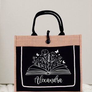 May include: A black and brown tote bag with a black pocket featuring a white outline of an open book with flowers and butterflies. The name "Alexandra" is written in white cursive below the book.