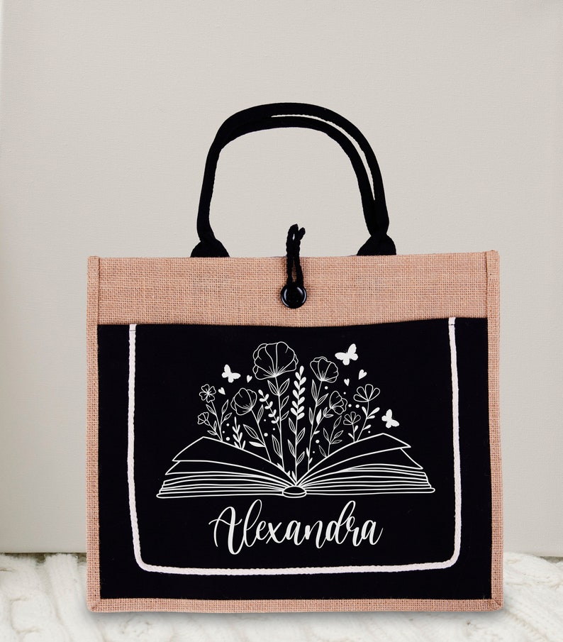 May include: A black and brown tote bag with a black pocket featuring a white outline of an open book with flowers and butterflies. The name "Alexandra" is written in white cursive below the book.