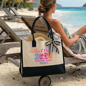 Pu&ograve; includere: Una borsa tote beige con manici neri e fondo nero, su una spiaggia. La borsa presenta il testo "Mother & Day 2026" in lettere colorate, insieme a "Seminyak Beach talitha". Un cappello di paglia e un fiocco a righe bianche e nere sono attaccati.