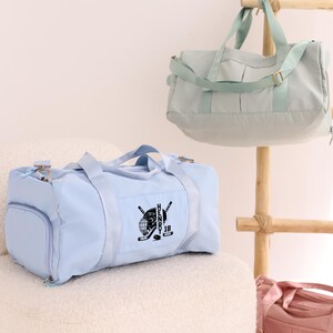May include: Two light blue duffel bags with black straps and zippers. One bag has a black hockey-themed design with the name "HENRY 18" on it. The bags are sitting on a white fuzzy surface.