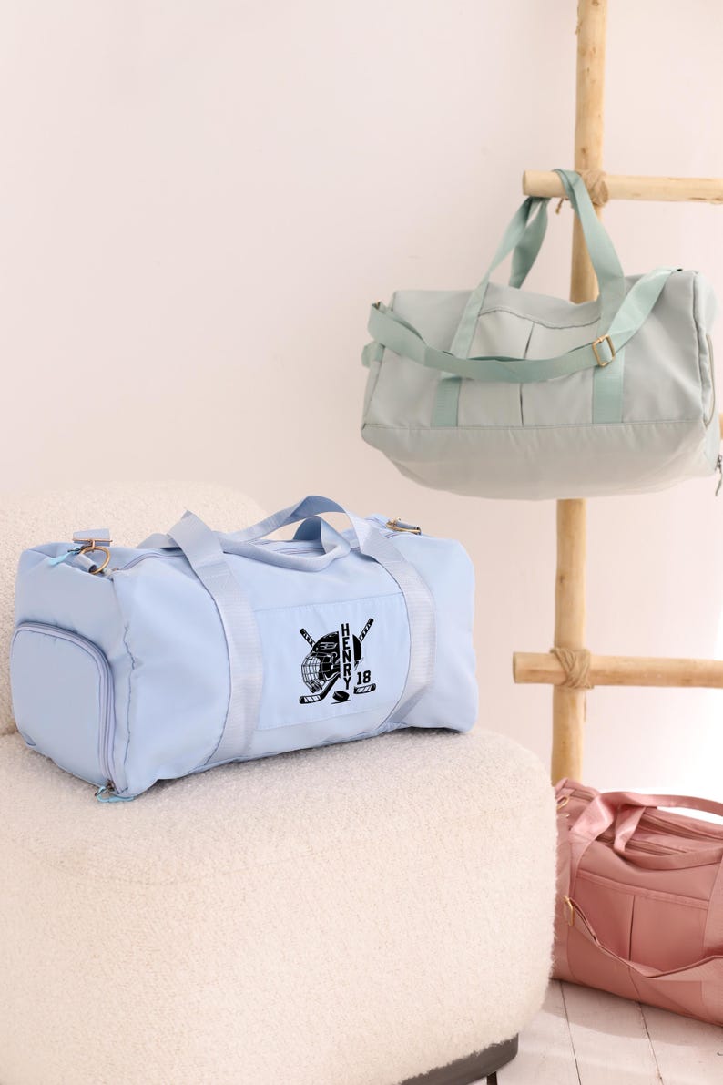 May include: Two light blue duffel bags with black straps and zippers. One bag has a black hockey-themed design with the name "HENRY 18" on it. The bags are sitting on a white fuzzy surface.