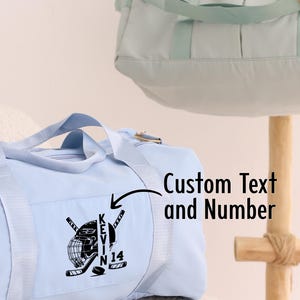 May include: Light blue duffel bag with a hockey helmet and sticks graphic and the text "KEVIN 14" embroidered on the front. Custom Text and Number.