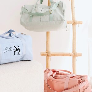 May include: Three pastel-coloured duffel bags: light blue, mint green, and pink. The light blue bag has the name "Elisa" and a gymnast silhouette printed on it. The bags are displayed near a wooden ladder.