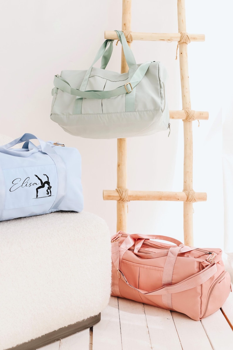 May include: Three pastel-coloured duffel bags: light blue, mint green, and pink. The light blue bag has the name "Elisa" and a gymnast silhouette printed on it. The bags are displayed near a wooden ladder.