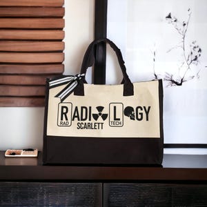 May include: A black and beige tote bag with the words "RADIOLOGY" printed on the front. The bag has a black and white ribbon tied to the handles. The words "RAD", "SCARLETT", "TECH" and a skull and crossbones symbol are printed on the bag.