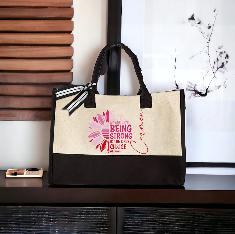 May include: A beige and black canvas tote bag with a pink and red sunflower design. The bag has a black handle and a black and white ribbon. The text on the bag reads "We don't know how strong we are until being strong is the only choice we have". The name "Carmen" is written on the bag in cursive.