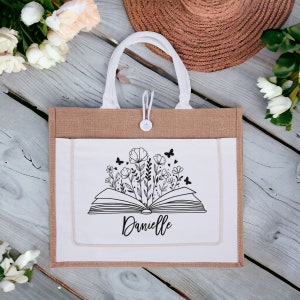 May include: A brown burlap tote bag with a white fabric pocket featuring a black and white illustration of an open book with flowers and butterflies. The text "Danielle" is printed below the book.