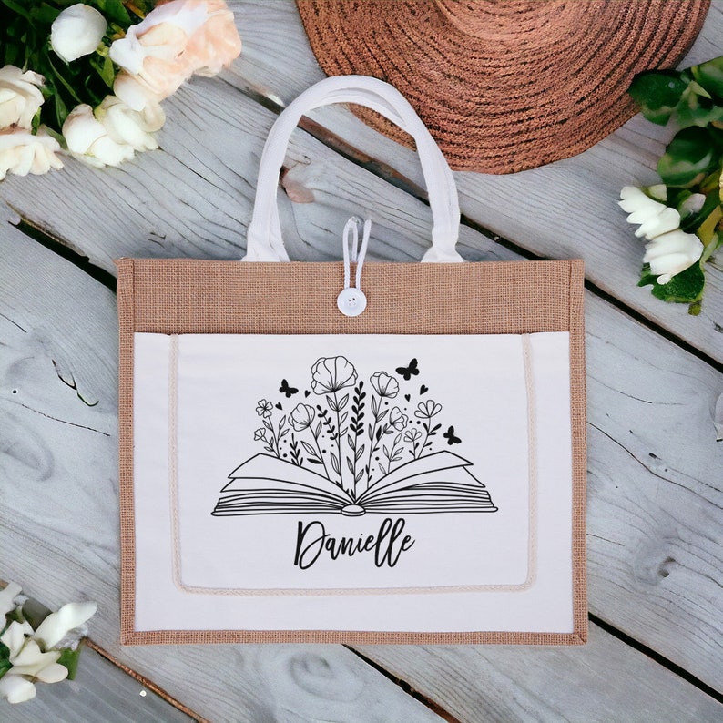 May include: A brown burlap tote bag with a white fabric pocket featuring a black and white illustration of an open book with flowers and butterflies. The text "Danielle" is printed below the book.