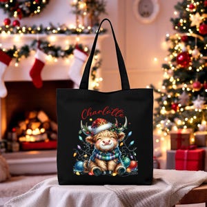 May include: Black tote bag featuring a cartoon Highland cow wearing a Santa hat and a plaid scarf, surrounded by Christmas lights and ornaments. The name "Charlotte" is written in red script above the cow. Festive holiday theme.