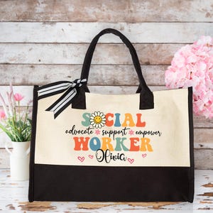 May include: A canvas tote bag with black handles and a black bottom panel. The bag features the words "SOCIAL WORKER" in colorful, retro-style lettering, along with the words "advocate support empower" and the name "Olivia". A black and white striped ribbon is tied to one of the handles.