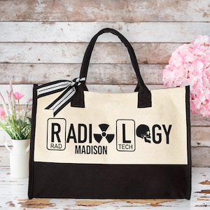 May include: A beige canvas tote bag with black handles and a black base. The bag has a black and white ribbon tied around the handles. The front of the bag has the words "RADIOLOGY" in black letters with a skull and crossbones symbol and the words "RAD" and "TECH" in smaller black letters. The words "MADISON" are printed in black letters below the word "RADIOLOGY".