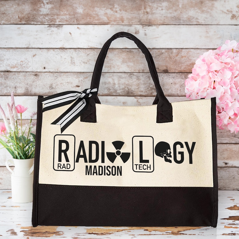 May include: A beige canvas tote bag with black handles and a black base. The bag has a black and white ribbon tied around the handles. The front of the bag has the words "RADIOLOGY" in black letters with a skull and crossbones symbol and the words "RAD" and "TECH" in smaller black letters. The words "MADISON" are printed in black letters below the word "RADIOLOGY".