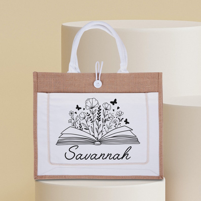 May include: A white tote bag with a brown burlap base. The bag has a white canvas panel with a black and white illustration of an open book with flowers and butterflies. The text "Savannah" is printed below the book.