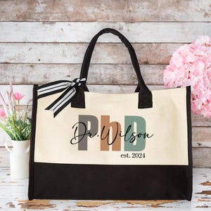 May include: A beige canvas tote bag with black handles and a black bottom. The bag has a black and white ribbon tied around the handles. The front of the bag has a colorful graphic that says "Dr. Wilson PhD est. 2024".