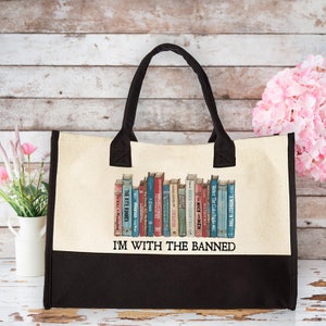 May include: A white canvas tote bag with black handles and a black bottom. The bag features a graphic of a bookshelf with a variety of books, including "The Kite Runner", "To Kill a Mockingbird", "The Giver", and "A Wrinkle in Time". The text "I'm With The Banned" is printed below the bookshelf graphic.