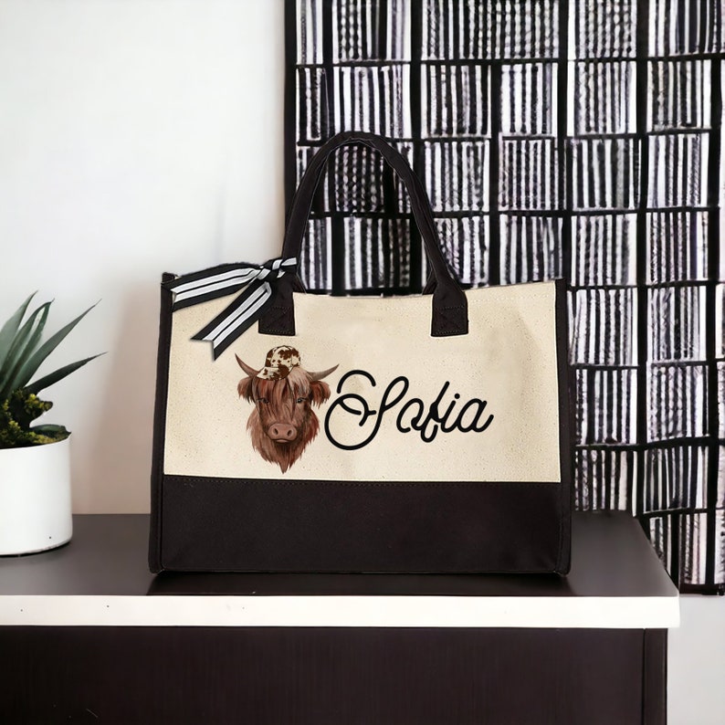 K&ouml;nnte beinhalten: Eine beige und braune Canvas-Tragetasche mit schwarzen Henkeln. Die Tasche hat eine braune Kuh mit einem Hut auf dem Kopf und den Namen "Sofia" in Kursivschrift auf der Vorderseite.