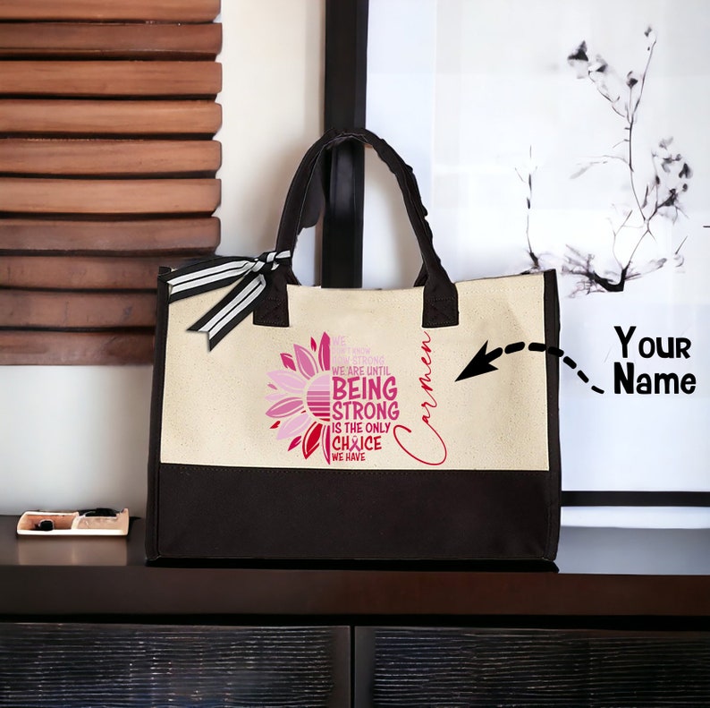 May include: A black and beige tote bag with a sunflower design and the text "We don't know how strong we are until being strong is the only choice we have". The bag has a black handle and a white ribbon. The text "Carmen" is written on the side of the bag. The text "Your Name" is written on a white background.