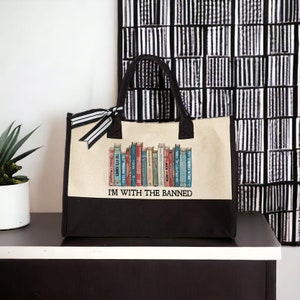 Peut inclure: Un sac fourre-tout en toile beige avec des anses noires et un fond noir. Le sac pr&eacute;sente un graphique d'une biblioth&egrave;que avec une vari&eacute;t&eacute; de livres color&eacute;s. Le texte "I'M WITH THE BANNED" est imprim&eacute; en noir sur le sac.