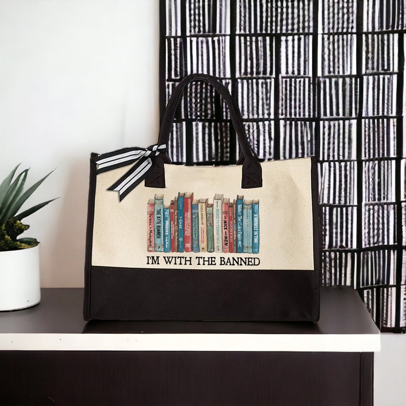 Peut inclure: Un sac fourre-tout en toile beige avec des anses noires et un fond noir. Le sac pr&eacute;sente un graphique d'une biblioth&egrave;que avec une vari&eacute;t&eacute; de livres color&eacute;s. Le texte "I'M WITH THE BANNED" est imprim&eacute; en noir sur le sac.