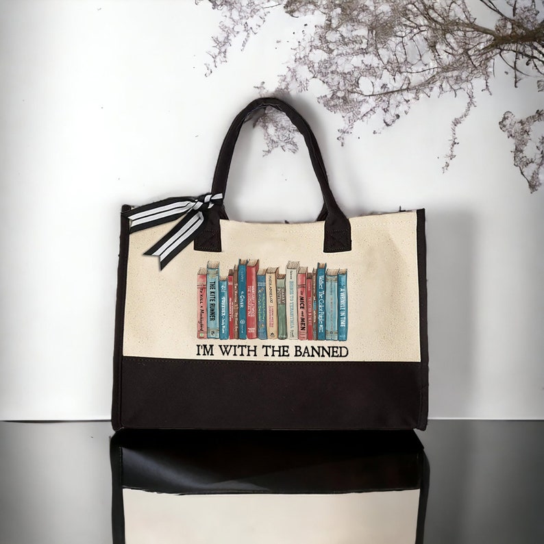 Peut inclure: Un sac fourre-tout en toile beige avec des anses noires et un fond noir. Le sac pr&eacute;sente un graphique d'une biblioth&egrave;que avec une vari&eacute;t&eacute; de livres. Le texte "I'M WITH THE BANNED" est imprim&eacute; sur le sac.