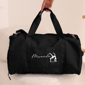 May include: Black duffel bag featuring a white gymnast graphic and the name "Miranda". The bag has two black handles and a front pocket. The bag is held by a person.
