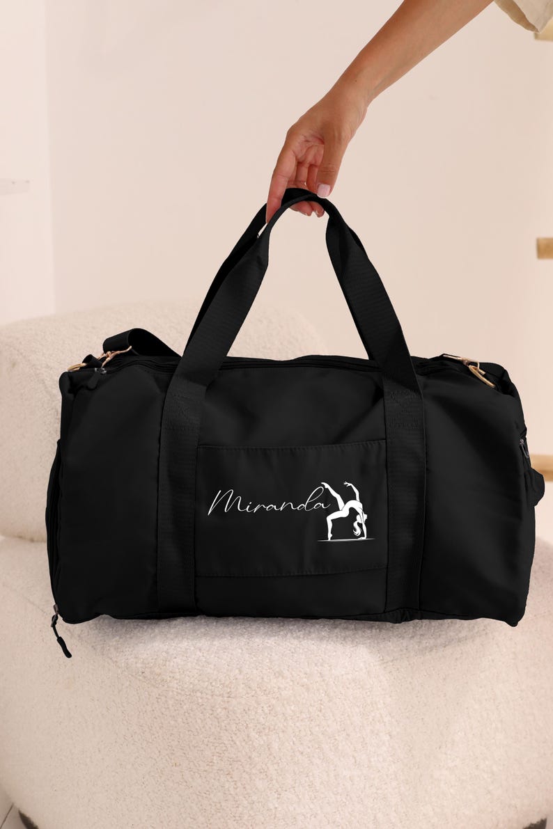 May include: Black duffel bag featuring a white gymnast graphic and the name "Miranda". The bag has two black handles and a front pocket. The bag is held by a person.