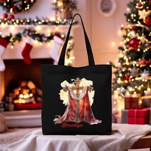 May include: Black tote bag featuring a detailed illustration of a cross with a red drape and a gold-colored cross. The word "Brooklyn" is written in red script at the bottom. The bag has black handles and is set against a festive background.