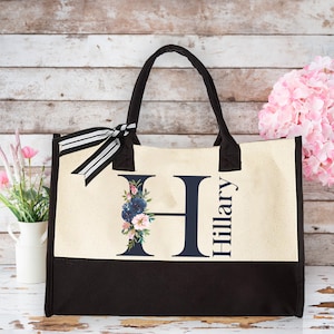 May include: A white canvas tote bag with black handles and a black bottom. The bag has a navy blue letter H with floral accents and the name "Hillary" printed on the front.