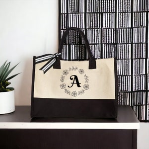 May include: A beige and black canvas tote bag with a black floral wreath and the letter "A" in the center.