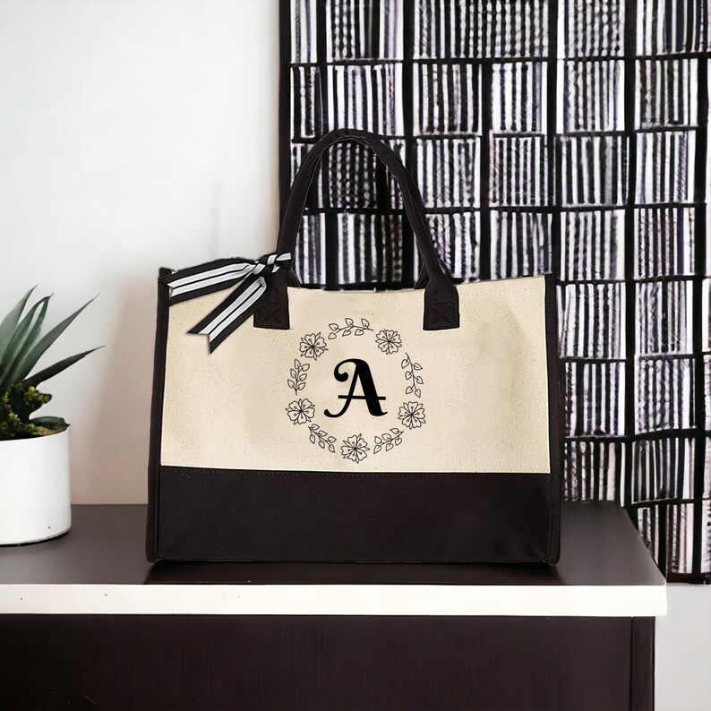 May include: A beige and black canvas tote bag with a black floral wreath and the letter "A" in the center.