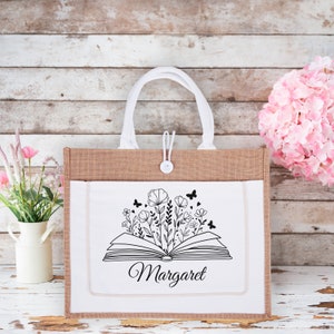 May include: A white tote bag with a brown burlap exterior. The bag has a white canvas front with a black and white illustration of an open book with flowers growing from it. The text "Margaret" is printed below the illustration.