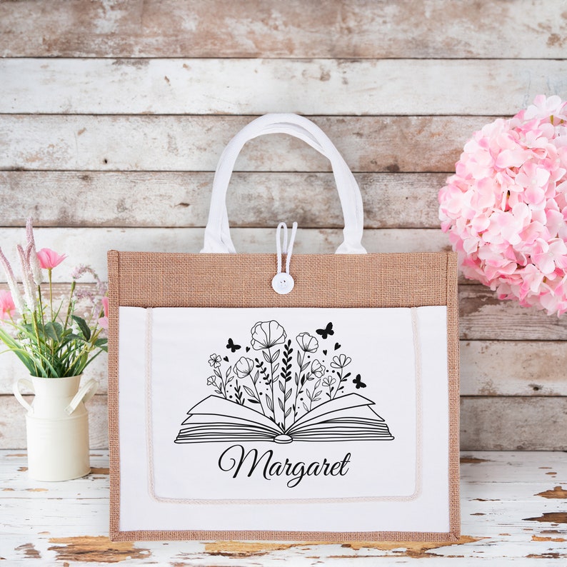 May include: A white tote bag with a brown burlap exterior. The bag has a white canvas front with a black and white illustration of an open book with flowers growing from it. The text "Margaret" is printed below the illustration.