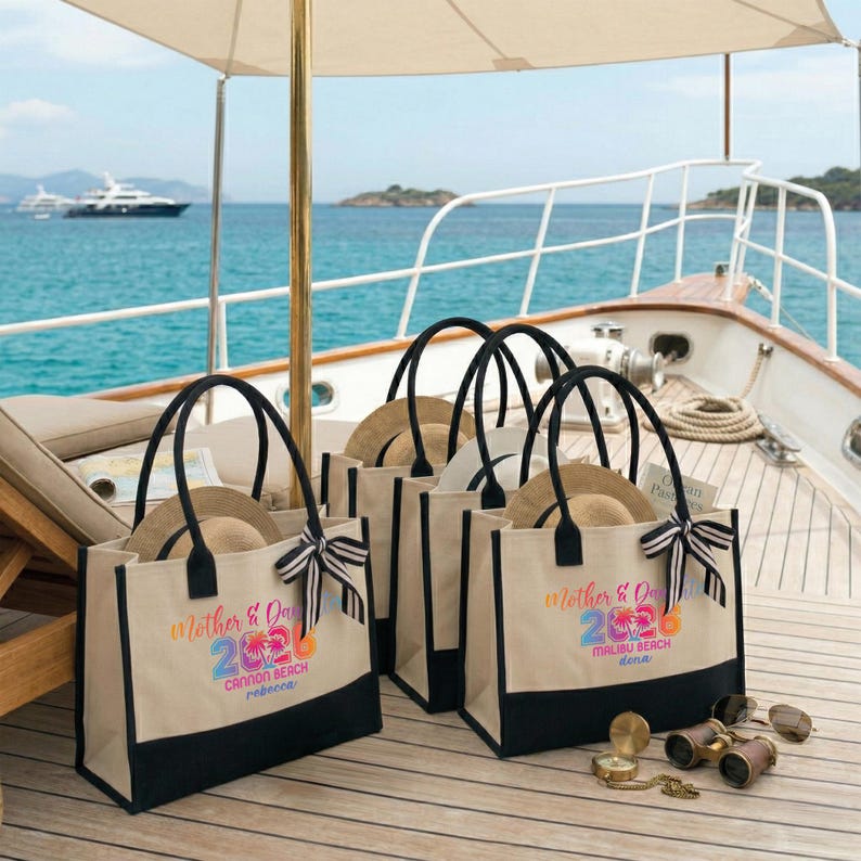 Pu&ograve; includere: Diverse borse tote in tela con manici e basi neri, ognuna contenente un cappello di paglia. Le borse presentano un design colorato "Mother & Day 2026" con nomi di localit&agrave;. Un fiocco a righe adorna una borsa. La scena &egrave; ambientata su una barca.