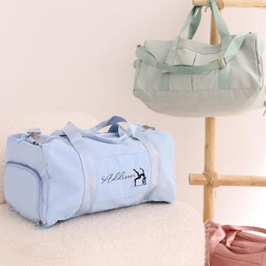 May include: Three pastel-coloured duffel bags: light blue, mint green, and pink. The light blue bag has the name "Addison" and a gymnast silhouette printed on it. The bags are displayed on a white chair and a wooden rack.