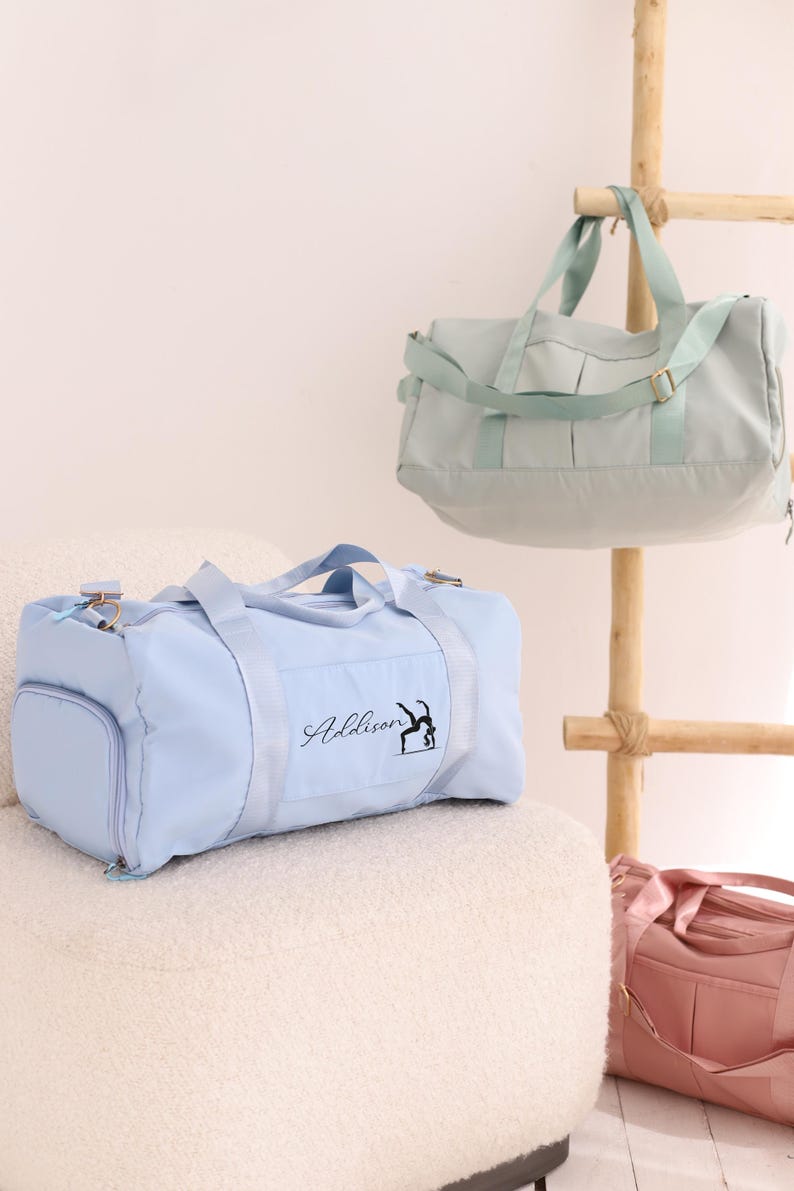May include: Three pastel-coloured duffel bags: light blue, mint green, and pink. The light blue bag has the name "Addison" and a gymnast silhouette printed on it. The bags are displayed on a white chair and a wooden rack.