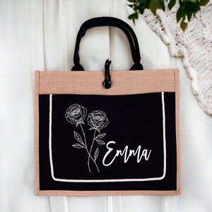 May include: A black and tan tote bag with a black fabric pocket featuring a white line drawing of two roses and the name "Emma".