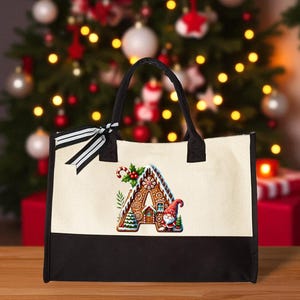 May include: A canvas tote bag with a black base and handles, featuring a Christmas-themed gingerbread letter "A" design. The letter is decorated with a gingerbread house, candy cane, and a Santa Claus figure. A black and white striped bow adorns the handle.
