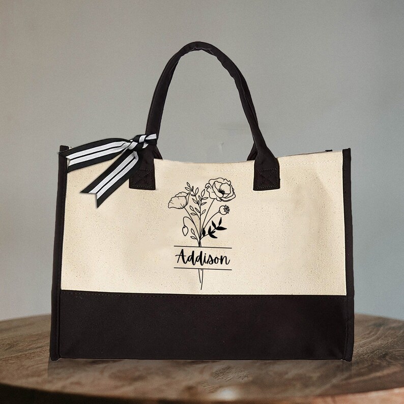 May include: A black and white tote bag with a floral design and the name "Addison" printed on the front. The bag has a black and white striped ribbon tied around the handles.