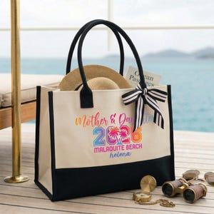 Pu&ograve; includere: Una borsa tote in tela color crema con manici neri e base nera. La borsa presenta la scritta "Mother & Daughter 2023 Malaquite Beach Helena" in colori arcobaleno. Un cappello di paglia e un libro sono all'interno. Un fiocco a righe bianche e nere &egrave; legato al manico.