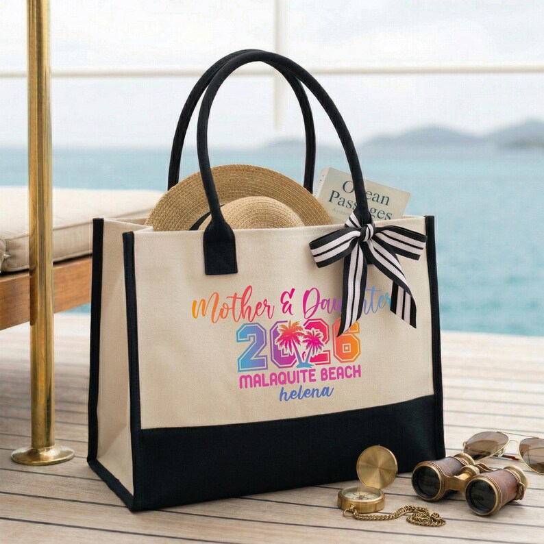 Pu&ograve; includere: Una borsa tote in tela color crema con manici neri e base nera. La borsa presenta la scritta "Mother & Daughter 2023 Malaquite Beach Helena" in colori arcobaleno. Un cappello di paglia e un libro sono all'interno. Un fiocco a righe bianche e nere &egrave; legato al manico.