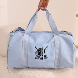 May include: Light blue duffel bag with a black hockey helmet, sticks, and puck design. The bag has a zippered pocket on the front and the text "DANIEL 10" is printed on the design.