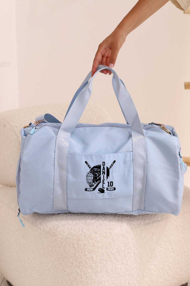 May include: Light blue duffel bag with a black hockey helmet, sticks, and puck design. The bag has a zippered pocket on the front and the text "DANIEL 10" is printed on the design.