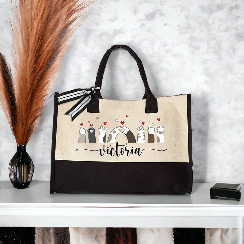 May include: A beige and black tote bag with a design of cat paws holding hearts and the name "victoria" printed on the front.